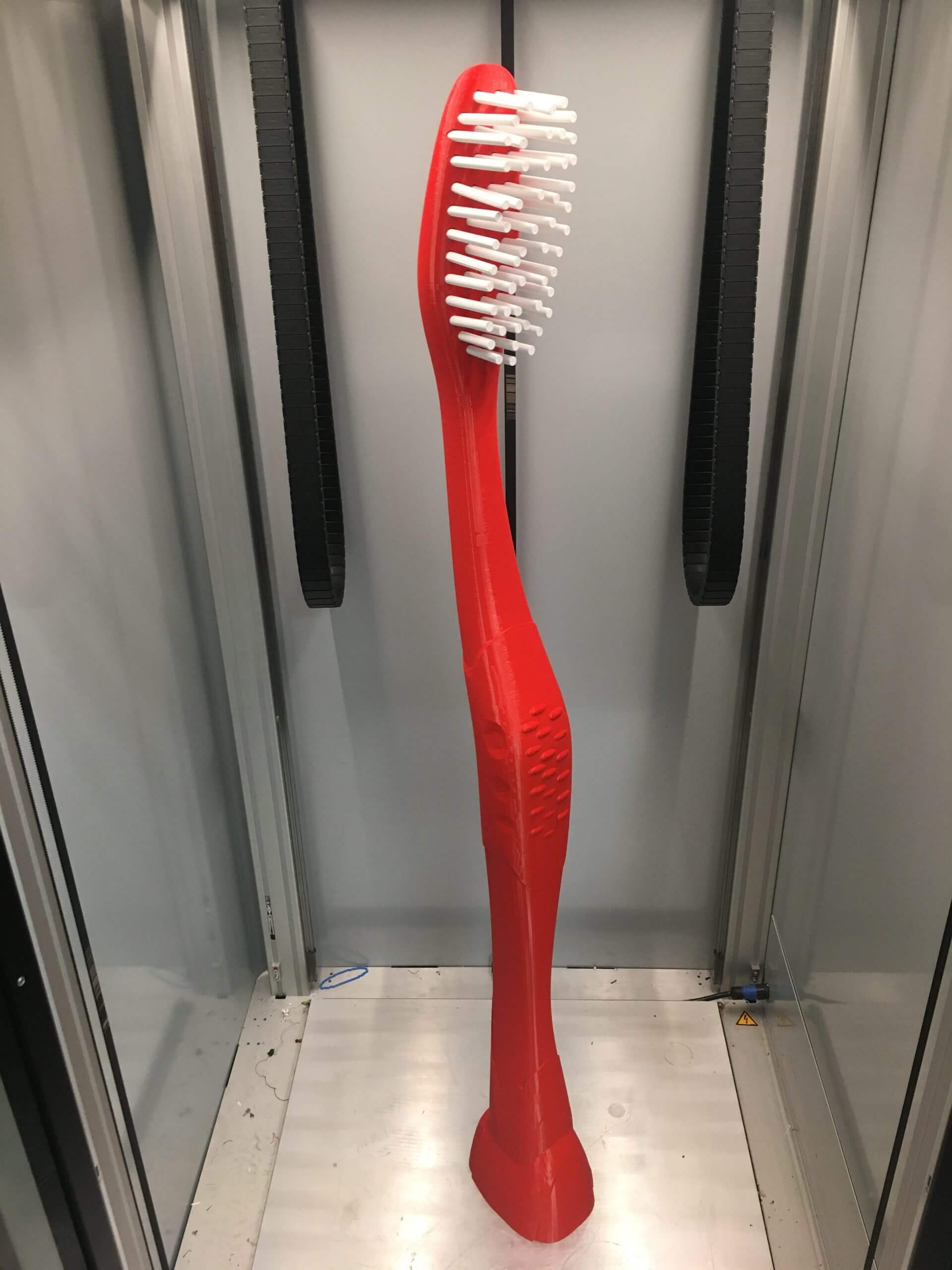 Brosse à dents rouge imprimée en 3D grand format avec poils blancs, présentée à l'intérieur d'une imprimante 3D industrielle