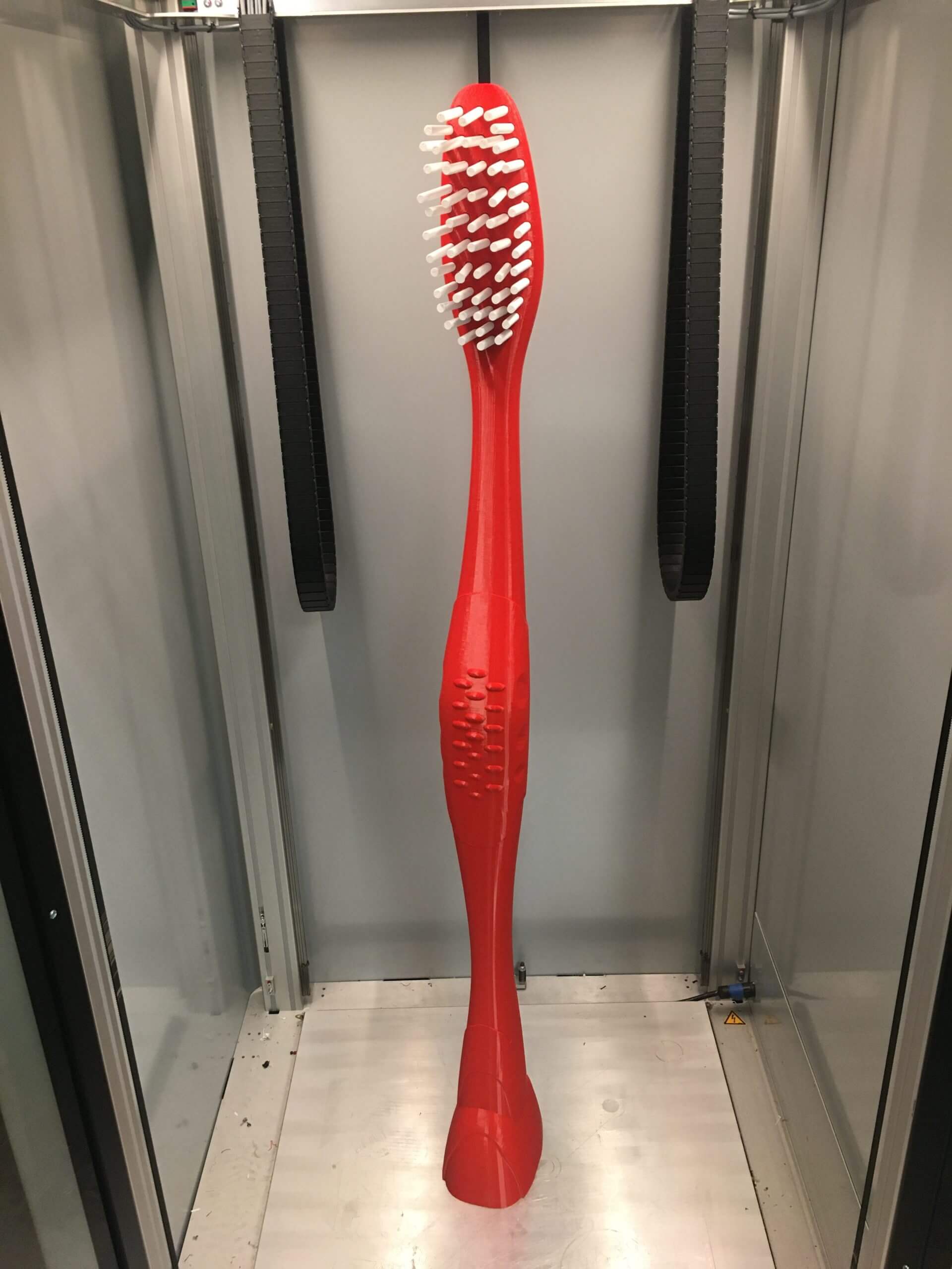 Brosse à dents rouge imprimée en 3D grand format avec poils blancs, présentée à l'intérieur d'une imprimante 3D industrielle