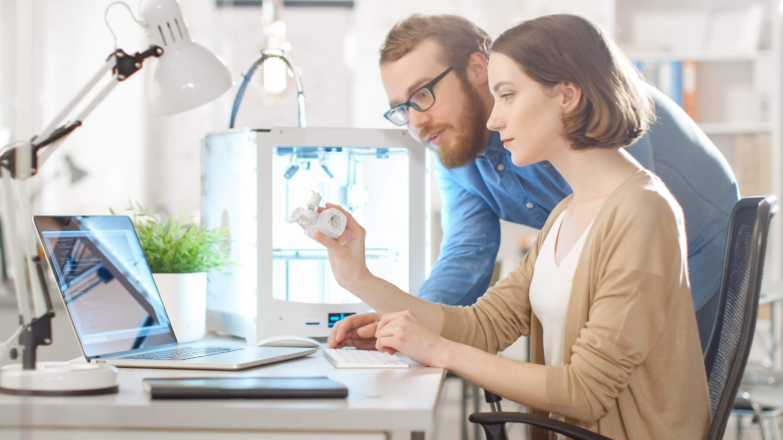 Deux personnes travaillant avec une imprimante 3D dans un bureau moderne, analysant une pièce imprimée en 3D.