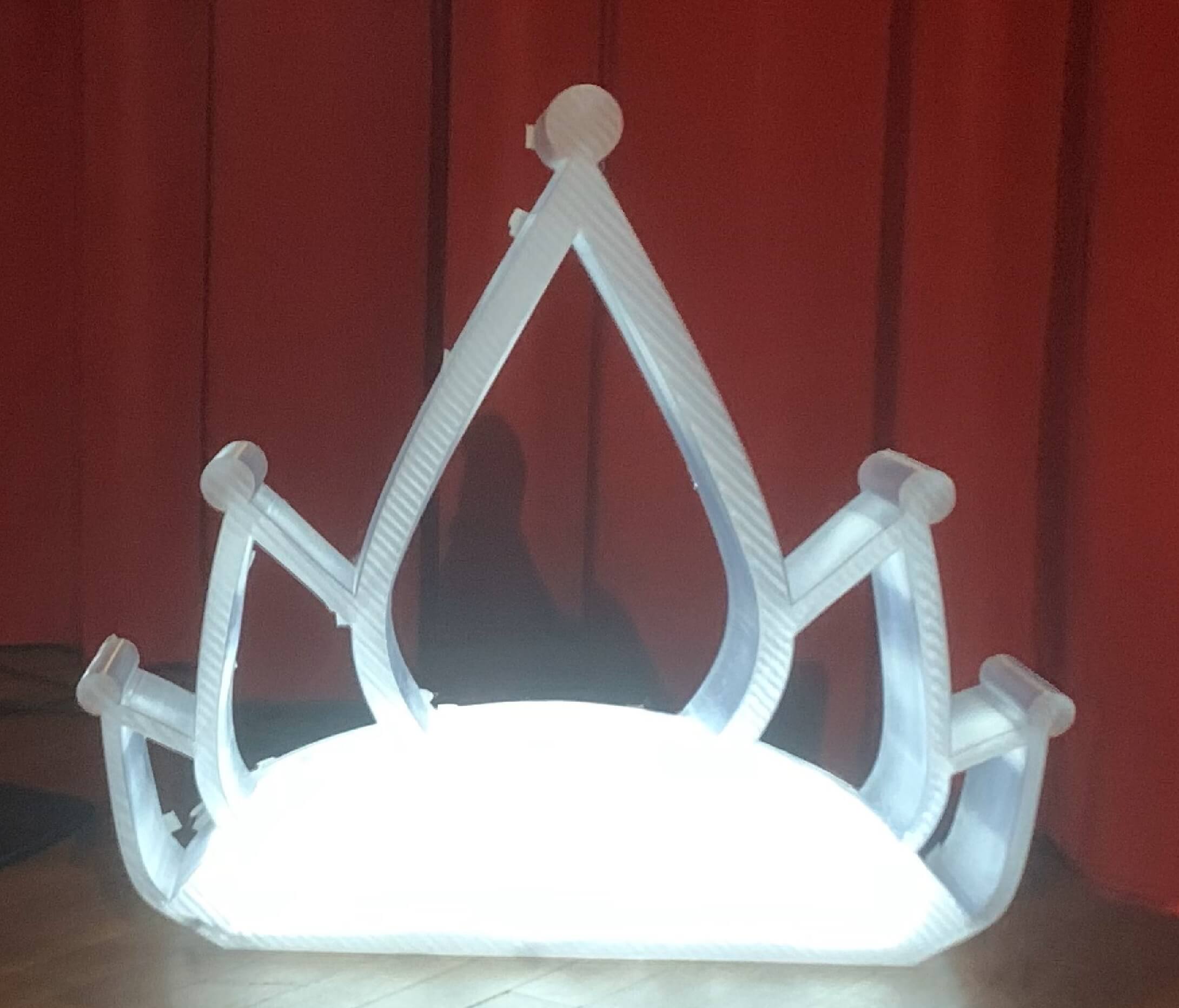 Couronne blanche imprimée en 3D grand format, éclairée par en dessous, posée sur un sol en bois avec un rideau rouge en arrière-plan