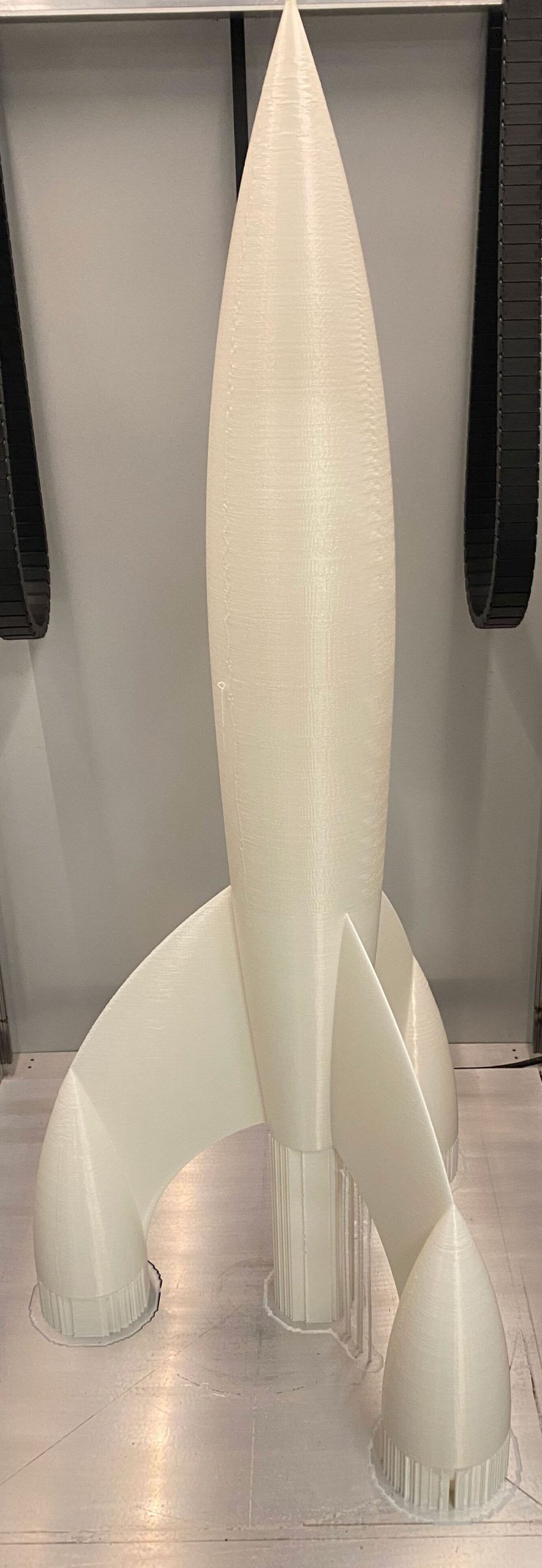 Fusée blanche imprimée en 3D grand format avec supports visibles, exposée dans un atelier d'impression 3D.